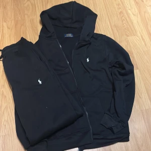 Raulph lauren tracksuit  - Svart tracksuit från Polo Ralph Lauren med dragkedja och huva. Klassisk vit broderad logga på bröstet, ribbade muddar och mjukt bomullsmaterial. Perfekt för chill dagar eller när du vill ha en clean och enkel look.