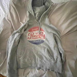 Grå Pepsi-Cola hoodie med tryck - Säljer en grå hoodie med klassiskt Pepsi-Cola tryck i rött, vitt och blått framtill. Tröjan har huva med snörning och en stor magficka. Perfekt för dig som gillar retrostil och vill ha något bekvämt och snyggt.