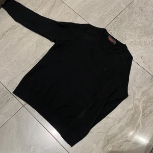 Svart merinoullströja från Morris - Klassisk svart stickad tröja från Morris i 100% merinoull. Tröjan har rund halsringning, diskret broderad logga på bröstet och ribbade muddar vid ärmslut och nederkant. Perfekt för lager-på-lager och stilren look.