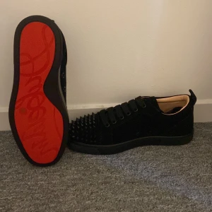 Svarta Louboutin sneakers med nitar - Säljer ett par svarta sneakers från Louboutin i mocka med ikonisk röd sula och svarta nitar över tån. Skorna har snörning och en låg siluett, perfekt för dig som vill sticka ut med en edgy och lyxig vibe.