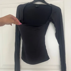 Svart långärmad topp från Gina Tricot - Svart långärmad topp från Gina Tricot med fyrkantig urringning. Tunn och stretchig bomullskvalitet som sitter tight mot kroppen. Perfekt för dig som gillar minimalistisk stil och vill ha en enkel men snygg topp till din garderob.
