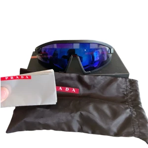 Prada sportiga svarta solglasögon - Säljer ett par feta svarta solglasögon från Prada med spegelblått, stort och sportigt visirglas. Ramen är svart och modellen har en futuristisk vibe. Kommer med originalpåse och putsduk med Prada-logga. Original pris 4900!