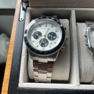 Faucon kronografklocka i silver - Stilren kronografklocka från Faucon med silverfärgad länk i metall och svart bezel. Urtavlan är vit med tre svarta subdials och tydliga indexmarkeringar. Klockan har tachymeter-skala runt urtavlan och datumvisning. Perfekt accessoar för dig som gillar sportig och klassisk stil.