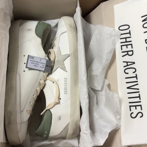 Golden Goose Superstar sneakers vit/grön - Golden Goose Superstar sneakers i vitt skinn med grå mockastjärna på sidan och grön häl. Grova vita snören och beige sula med retrovibbar. Snygg kontrast mellan material och färger, perfekt för dig som gillar streetstyle och unika detaljer.