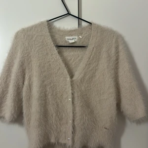 Beige fluffig kofta från Holly & White - Supermjuk och fluffig beige kofta från Holly & White med korta ärmar och v-ringad hals. Koftan har pärlknappar framtill och en croppad passform som ger en trendig look. Perfekt för lager-på-lager under kyliga dagar.