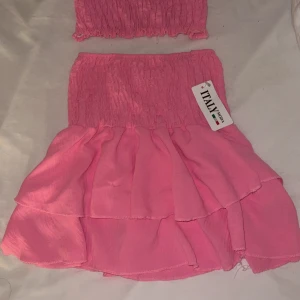 Rosa set från Italy Moda - Supersöt rosa kortkjol med volanger och top  från Italy Moda. Kjolen har smockad midja som ger en bekväm och snygg passform. Perfekt för dig som vill ha en feminin och trendig look. Tunt och lätt material som passar bra till varma dagar. Hela settet för 80kr
