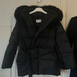 Svart dunjacka med päls från FUR Stockholm - Säljer en svart dunjacka från FUR Stockholm med fluffig päls runt huvan. Jackan har bälte i midjan, quiltad design och långa ärmar. Perfekt för kalla dagar med sin tjocka och mysiga känsla. Snygg och stilren look med detaljer som knappar och dragkedja. (Har lite fläckar vid luvan och lappen är av)