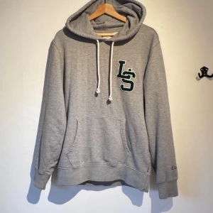 Grå hoodie från Levis med LS-tryck - Grå hoodie från Levis med broderat LS-tryck i grönt och vitt på bröstet. Tröjan har huva med snörning, magficka och ribbade muddar vid ärmar och nederkant. Perfekt för chill dagar och enkel att matcha med jeans eller joggers.