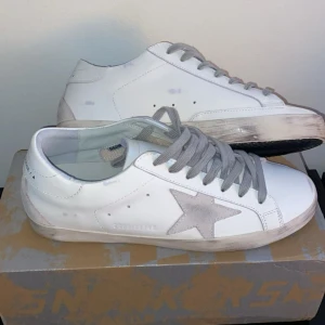 Golden Goose  - Golden Goose sneakers i vitt skinn med grå mockadetaljer och ikonisk stjärna på sidan. Skorna har grå snörning, platt sula och rund tå. Snyggt slitna detaljer på sulan för en cool, avslappnad look. Kommer med originalpåse och låda. Säljer då dem var lite små för mig, jag har 43 och storleken är lite mindre så passar perfekt på 42,5 och 42🌟 tveka inte att höra av dig vid fler frågor eller bilder🔥
