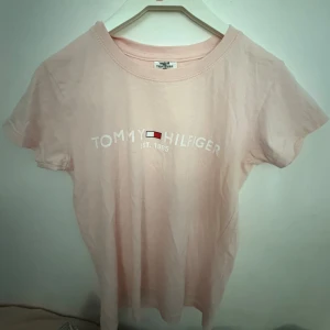 Ljusrosa t-shirt med Tommy Hilfiger-tryck - Ljusrosa t-shirt i bomull med klassiskt Tommy Hilfiger-tryck och logga framtill. Modellen är kortärmad med rund halsringning och har en clean, enkel look som passar perfekt till jeans eller shorts.