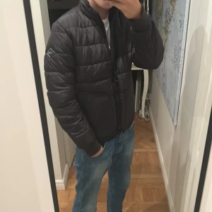 Svart pufferjacka från Replay - Säljer en svart pufferjacka från Replay med diskret logga på ärmen. Jackan har quiltad design, dragkedja framtill och hög krage. Perfekt för kyliga dagar och ger en clean, modern look. Materialet är syntet och jackan har lång ärm. Använd en höst säsong ser helt ny ut 