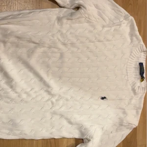 Vit kabelstickad tröja Polo Ralph Lauren - Kabelstickad vit tröja från Polo Ralph Lauren med långa ärmar och rund hals. Klassisk design med broderad svart logga på bröstet. Perfekt för dig som gillar preppy stil och vill ha något stilrent och tidlöst.