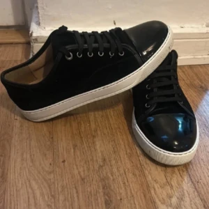 Svarta Lanvins med gläns där framme - Säljer ett par svarta sneakers från Lanvin med vit sula och snygg lackad tå. Skorna har snörning och är tillverkade i mocka och skinn, vilket ger en lyxig känsla. Perfekta för dig som vill ha en stilren och trendig look.