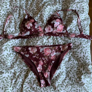 Blommig vinröd bikini från H&M - Supersöt bikini från H&M i vinröd färg med blommigt mönster i rosa och vitt. Klassisk trekantsmodell upptill med knytband och matchande bikinitrosa. Oanvänd.