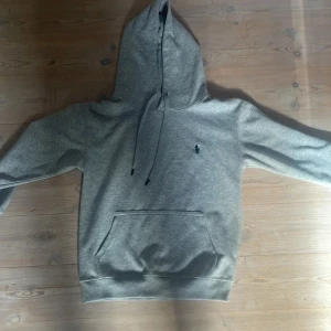 Grå hoodie från Polo Ralph Lauren - Snygg grå hoodie från Polo Ralph Lauren med klassisk svart broderad logga på bröstet. Tröjan har huva med snörning, stor magficka och är tillverkad i mjuk bomull. Perfekt för en chill och stilren look. Passar om du är 155-165