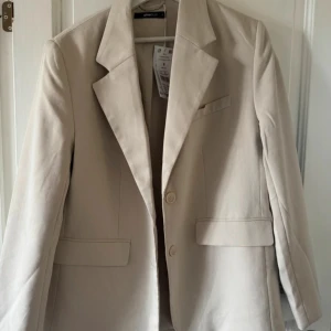 Oversized beige kavaj från Gina Tricot - Stilren oversized kavaj i beige från Gina Tricot. Klassisk design med två knappar framtill, breda slag och fickor med lock. Perfekt för dig som gillar en avslappnad men ändå dressad look. Färgen är ljus och passar till det mesta.