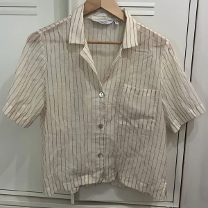 &Other Stories Randig kortärmad skjorta  - Snygg kortärmad skjorta från & Other Stories med tunna vertikala ränder i beige och vitt. Skjortan har klassisk krage, bröstficka och knäppning framtill.