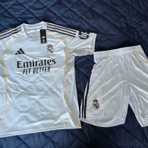 Real Madrid matchtröja Adidas vit - Säljer en officiell Real Madrid fotbollströja från Adidas i vitt med svarta detaljer och gula inslag. Tröjan har klubbmärke och sponsortryck framtill, samt klassiska Adidas-ränder på axlarna. Tillverkad i lätt funktionsmaterial.