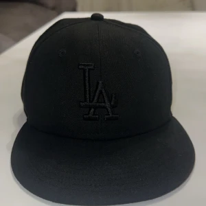 Svart LA-keps från New Era 59FIFTY - Säljer en svart New Era 59FIFTY keps med LA-logga broderad framtill. Klassisk flat peak och broderade ventilationshål. Kepsen är helsvart, även detaljerna och loggorna. Perfekt för dig som gillar stilrena accessoarer och streetwear.