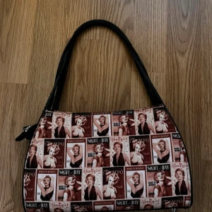 Retro Marilyn Monroe handväska - Cool handväska med retroinspirerat Marilyn Monroe-tryck i bruna och beige toner. Väskan har svart handtag och detaljer, samt dragkedja upptill. Perfekt accessoar för dig som gillar vintagevibbar och unika prints.
