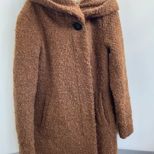 Brun teddyjacka med huva från VILA - Mysig brun teddyjacka från VILA med stor huva och knäppning framtill. Jackan har en fluffig, teddy-liknande yta och två fickor på sidorna. Perfekt för kyliga dagar när du vill ha både stil och komfort.