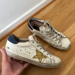Golden Goose superstars med guldglittrig stjärna - Golden Goose Superstars sneakers, Vita med guldglittrig stjärna med mörkblått vid hälen. Skorna är andvända i gott skick. Storlek 39