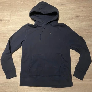 Mörkblå hoodie från J.Lindeberg - Säljer en stilren mörkblå hoodie från J.Lindeberg med broderad logga på bröstet. Tröjan har huva med snörning, magficka och långa ärmar. Perfekt för dig som gillar en clean och enkel look. Efter lite kärlek kan denna se ut som ny
