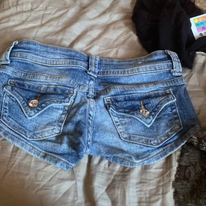 Blå jeansshorts från Bershka - Säljer ett par blå jeansshorts från Bershka i klassisk denim med låg midja. Shortsen har två bakfickor med snygga knappar, dubbla knappar fram och dragkedja. Passformen är tight och de har en cool, ungdomlig vibe. Perfekta för varma dagar!
