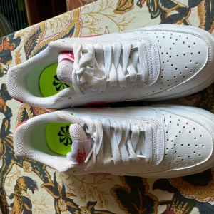 Nike sneakers vit/rosa strl 38 - Snygga vita Nike sneakers med rosa detaljer och klassisk swoosh på sidan. Skorna har snörning, perforerad tå och bekväm platt sula. Tillverkade i syntet och skinnliknande material. Perfekta för dig som gillar en fräsch och sportig look.