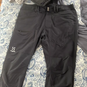 Svarta täckbyxor från Haglöfs - Svarta täckbyxor från Haglöfs med dragkedjor vid fickorna och logga på vänster ben. Byxorna har bälteshällor och knappstängning i midjan. Perfekta för utomhusaktiviteter när det är kallt.