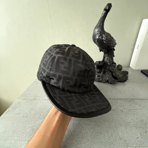 Svart Fendi keps med FF-mönster - Snygg svart keps från Fendi med ikoniskt FF-mönster över hela kepsen. Justerbar baktill med metallspänne och Fendi Roma-logga. 