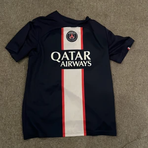 PSG matchtröja Messi #30 M - Säljer en officiell PSG fotbollströja i marinblått med vit och röd rand mitt på, Qatar Airways-tryck och Messi #30 på ryggen. Tröjan är kortärmad och tillverkad i lätt, andasvänligt material. Perfekt för dig som älskar fotboll och vill bära din favoritspelare.