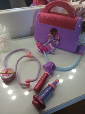 Doktorsväska i plast, lila och rosa - Säljer en söt doktorsväska i plast från Doc McStuffins. Setet innehåller lila och rosa leksaksstetoskop, spruta, termometer och andra doktorsredskap, allt med glitterdetaljer. Perfekt för rollekar och lekar hemma.
