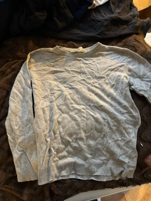 Ljusgrå stickad tröja från H&M - En enkel och stilren ljusgrå stickad tröja från H&M. Tröjan har rund halsringning, ribbade muddar vid ärmslut och nederkant samt en avslappnad passform. Perfekt att matcha med jeans eller kjol för en clean look.