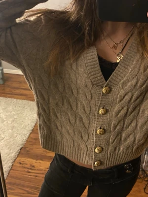 Beige kabelstickad kofta med guldknappar - Snygg beige kabelstickad kofta med djup V-ringning och dekorativa guldknappar framtill. Kortare modell med ribbade muddar och mjukt material som ger en chill vibe. Perfekt att slänga över ett linne eller topp.