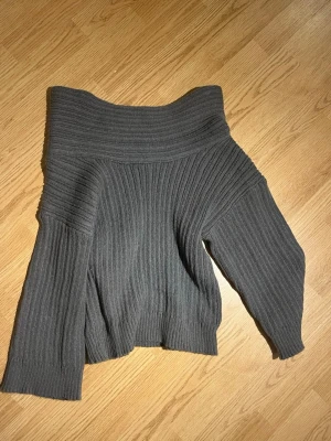 Mörkgrå ribbstickad tröja - Trendig mörkgrå stickad tröja med bred ribbstickning och off shoulder-design. Tröjan har långa ärmar och en bred, vikt krage som ger en cool och avslappnad look. Perfekt för lager på lager och passar till både jeans och kjol.