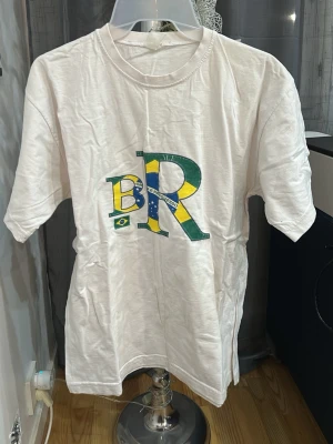 Vit BR t-shirt med Brasilien-tryck - Vit t-shirt med stort BR-tryck i brasilianska flaggans färger på bröstet. Klassisk rund hals och korta ärmar. Perfekt för dig som vill visa kärlek till Brasilien på ett stilrent sätt.