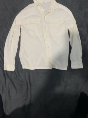 Vit skjorta med fickor framtill - Klassisk vit skjorta med två bröstfickor och knäppning framtill. Skjortan har lång ärm och en rak passform, perfekt för en stilren look. Tillverkad i ett mjukt bomullstyg som känns skönt mot huden.