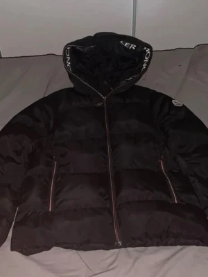 Svart Moncler dunjacka med huva - Snygg svart dunjacka från Moncler med stor huva och tydlig logga på ärmen. Jackan har dragkedja framtill och två fickor med dragkedja. Moncler-tryck på huva och insida ger en exklusiv känsla. Perfekt för kalla dagar och riktigt street.