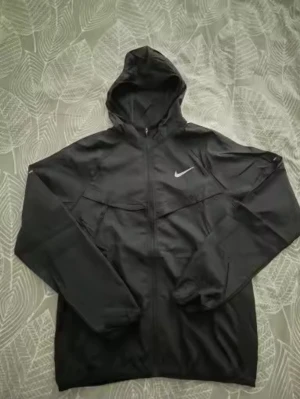 Svart Nike React windbreaker med huva - Svart  windbreaker från Nike React med klassisk vit logga på bröstet. Jackan har huva, hel dragkedja framtill och långa ärmar. Tillverkad i lätt och smidigt syntetmaterial, perfekt för blåsiga dagar. Enkel och stilren design som passar till många outfits.