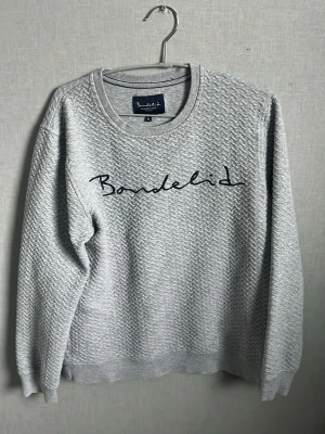 Grå sweatshirt från Bondelid - Säljer en grå sweatshirt från Bondelid med broderad logga på bröstet. Tröjan har rund halsringning, lång ärm och ett diskret fiskbensmönster i tyget. Perfekt för en avslappnad och stilren look.