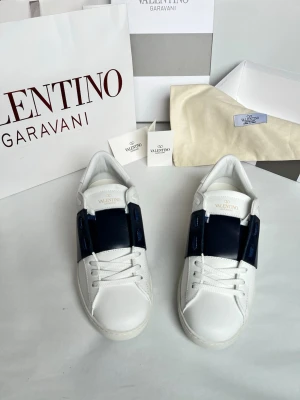 Valentino Garavani vita sneakers - Snygga vita sneakers från Valentino Garavani med en bred mörkblå detalj över snörningen. Skorna har rund tå, platt sula och är tillverkade i skinn. Perfekta för dig som vill ha en clean men ändå unik look.