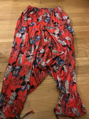 Röda blommiga shorts med snörning - Färgstarka röda shorts med blommigt mönster i blått, vitt och lila. Shortsen har elastisk midja och snörning vid bensluten för en avslappnad look. Perfekta för varma dagar och ger en riktigt somrig vibe.
