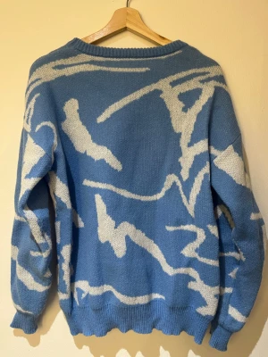 Blå/vit stickad tröja - Cool stickad tröja i blått med vitt abstrakt mönster. Tröjan har rund halsringning, långa ärmar och ribbade muddar vid ärmslut och nederkant. Perfekt för dig som gillar unika plagg med streetkänsla.