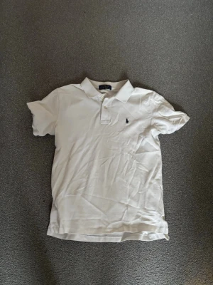 Vit pikétröja från Ralph Lauren - Klassisk vit pikétröja från Ralph Lauren med korta ärmar och krage. Tröjan har knappar vid halsen och den ikoniska broderade loggan på bröstet. Perfekt för en stilren och avslappnad look. Pris kan diskuteras!