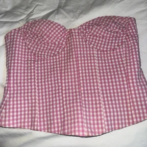 Rosa rutigt korsett från Zara - Supersöt korsett från Zara i rosa och vitt rutigt mönster. Modellen har markerade kupor och vertikala sömmar för en snygg siluett. Stängs med hakar på sidan. Perfekt för att styla lager på lager eller som statement-plagg.