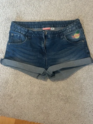 Mörkblå jeansshorts från Pepperts - Snygga mörkblå jeansshorts från Pepperts med uppvikta benslut och en liten broderad ananas på fickan. Klassisk femficksmodell med dragkedja och knapp. Perfekta för varma dagar och enkel att matcha med olika toppar.