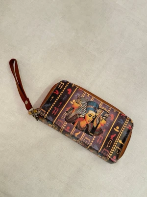 Färgstark egyptisk plånbok med tryck från Egyptiern  - Unik plånbok med färgstarkt egyptiskt motiv och hieroglyfer. Köpt från egyptiern. Plånboken har dragkedja och en brun handledsrem. Tillverkad i syntetmaterial med detaljer i brunt, blått, rött och gult. Perfekt för dig som gillar statement-accessoarer.