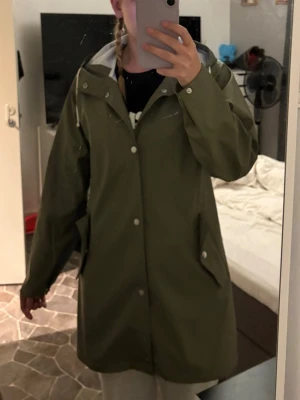 Regnkappa  - Snygg olivgrön regnjacka med huva och tryckknappar framtill. Jackan har två stora fickor med lock och en rak, loose passform. Perfekt för regniga dagar och enkel att matcha med olika outfits.