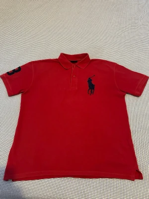 Röd pikétröja från Ralph Lauren - Klassisk röd pikétröja från Ralph Lauren med stor marinblå logga på bröstet och siffran 3 broderad på ena ärmen. Tröjan har krage, knappar framtill och är kortärmad. Perfekt för en sportig och stilren look.